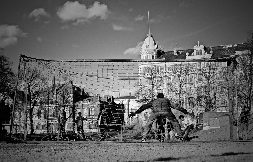 Framed by towering city buildings, the goalkeeper steadies himself before the enormous net, preparing for the high-stakes penalty shot.  "Goalie" by laureltopias is licensed under CC BY 2.0.